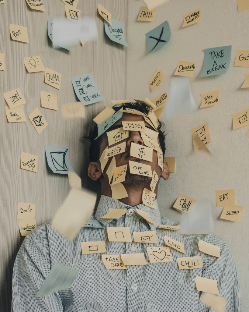 man leaning up against a wall with many postits covering his face and wall with tasks written on each.