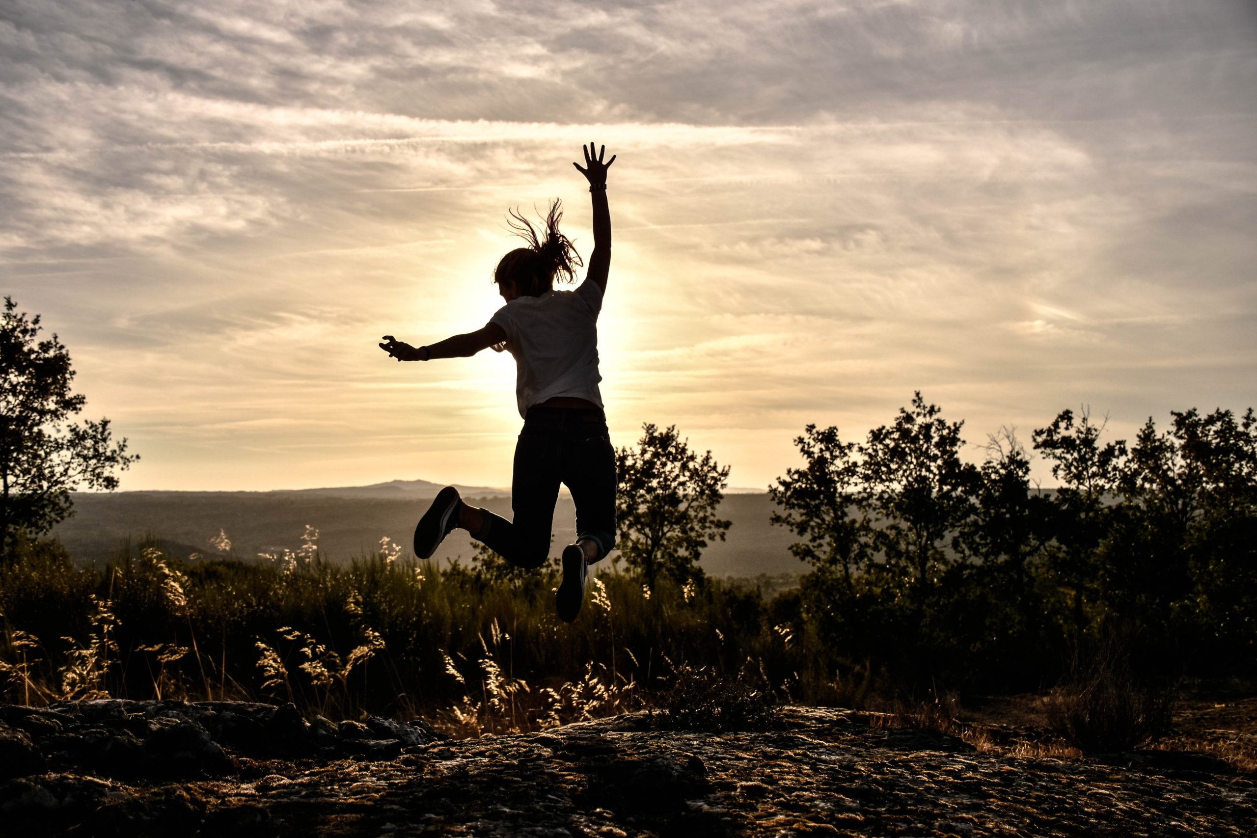 person jumping in excitement in the air in front of the sunset. Some trees and hills noticed.