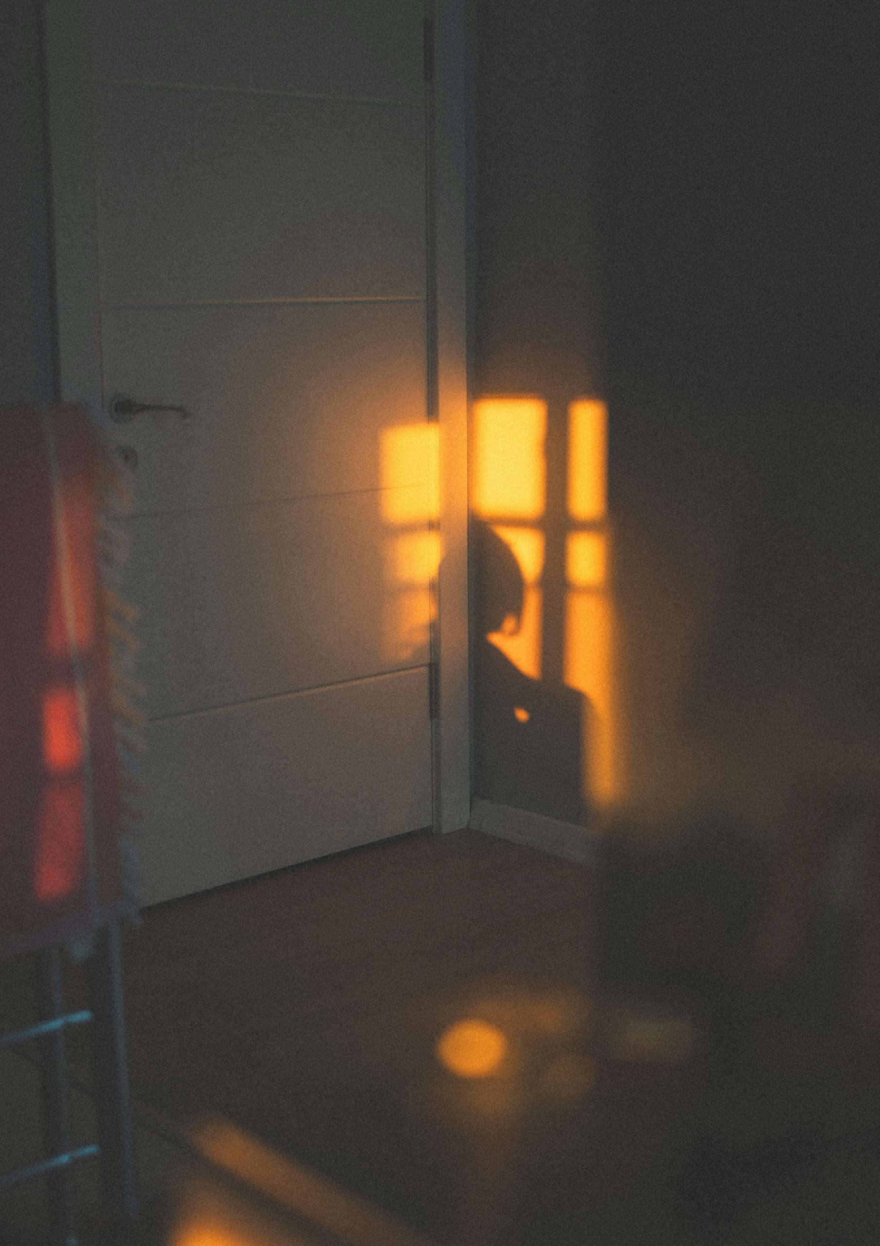 DARK BEDROOM DOOR AND WALL WITH YELLOW SUNLIGHT SHINING ON THE WALL SHOWING A SHADOW OF A HUMAN BODY SITTING WITH KNEES CURLED UP AND HEAD HANGING DOWN