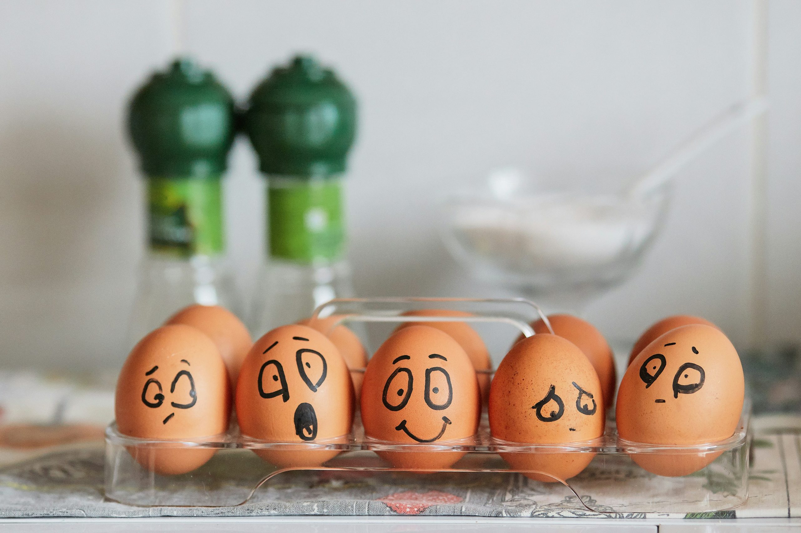 brown eggs in a clear carton on the counter. each egg has a different emotional face on it.
